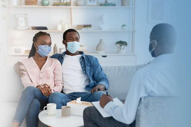 家庭治疗师与坐在办公室沙发上戴着防护面具的黑人夫妇交谈
