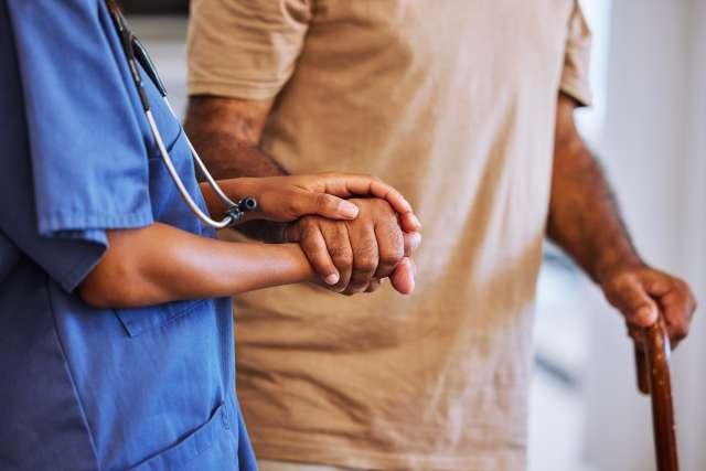 A caregiver holds a patient's hand, helping the patient to walk.